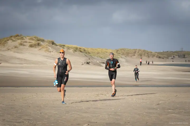 Twee deelnemers in wetsuits lopen over het strand naar de camera, een van hen met oranje badmuts.