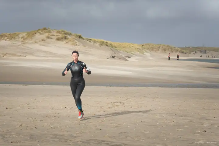 Een loopster in wetsuit steekt beide duimen op terwijl ze over het strand komt aanrennen.
