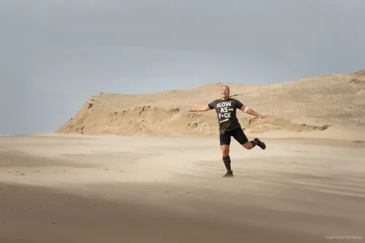 Een deelnemer springt uitgelaten op het strand met gespreide armen, een zandduin op de achtergrond.