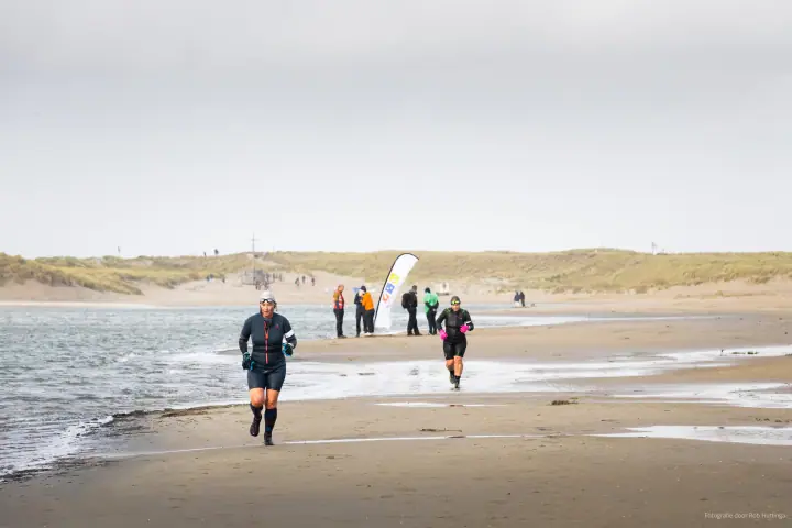 Twee lopers volgen de waterlijn, met toeschouwers en een beachvlag verderop.