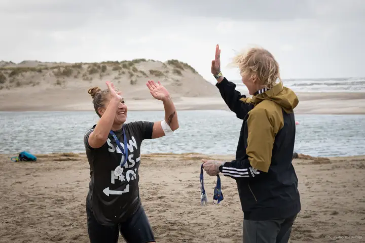 Een finisher en een vrijwilliger met medailles in haar hand geven elkaar een high five aan het water.