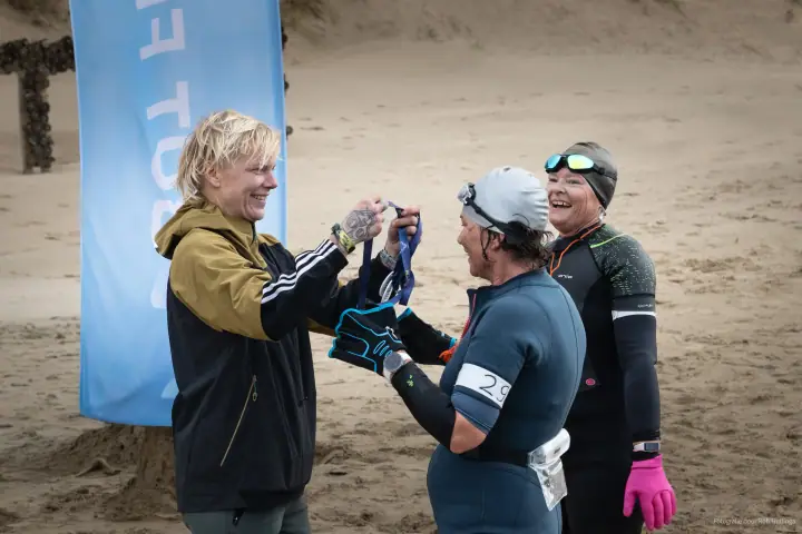 Een vrijwilliger hangt een medaille om bij een lachende finisher in badmuts, een tweede finisher lacht mee.