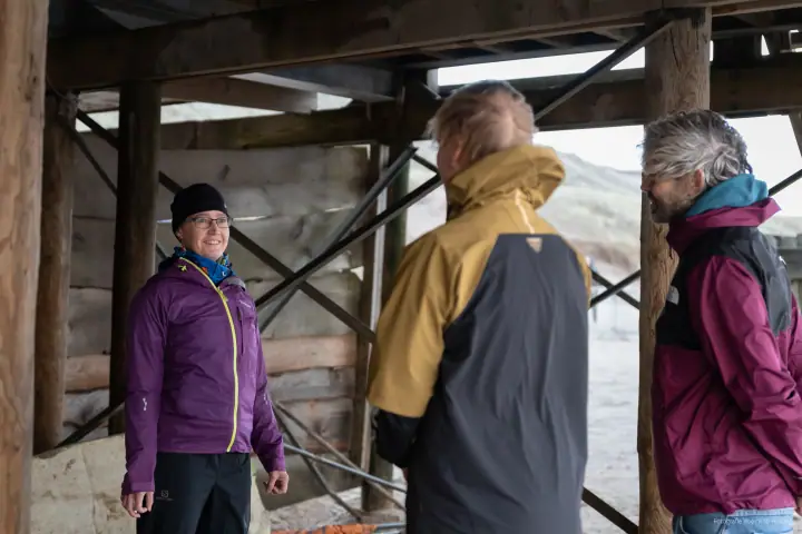 Drie mensen in regenjassen staan te praten onder het houten strandpaviljoen.