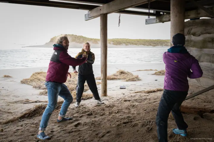 Onder het strandpaviljoen bewegen drie mensen uitbundig met hun armen, de zee op de achtergrond.