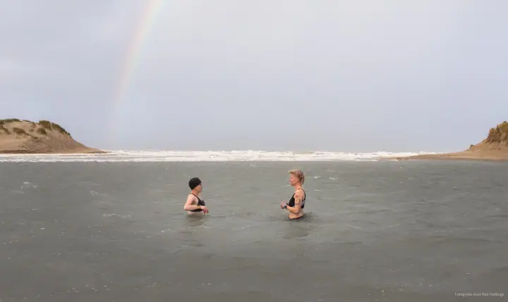 Twee vrouwen staan tot hun middel in het water, met een regenboog boven de duinen.