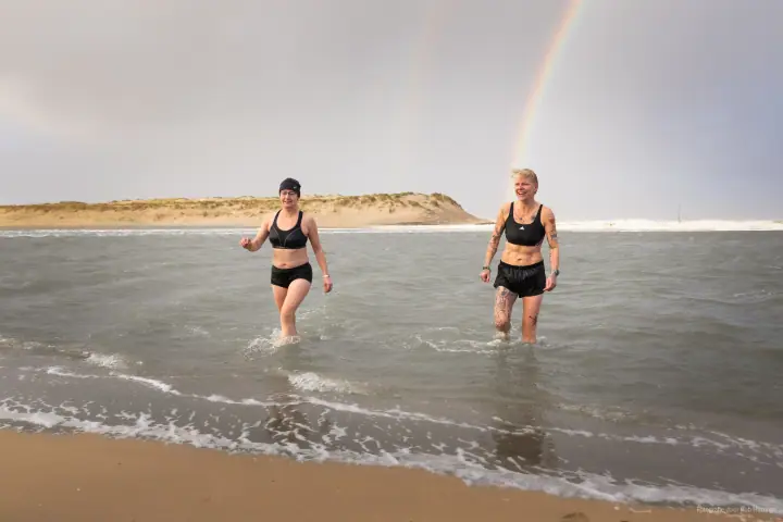 Twee vrouwen in sportkleding lopen lachend het water uit, een regenboog boven de duin.