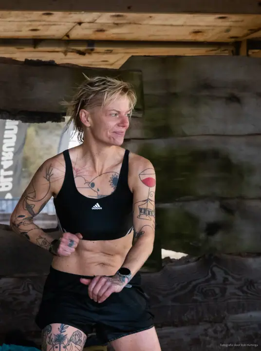 Portret van een getatoeëerde deelnemer in sporttop onder het houten paviljoen, haar haar nog nat.