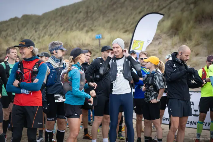 Trailrunners met hardloopvesten verzamelen zich bij de start, voor een duin vol helmgras.