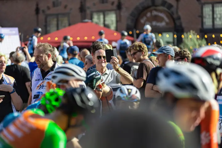 Publiek op de Grote Markt filmt de renners met de telefoon, met De Waag op de achtergrond