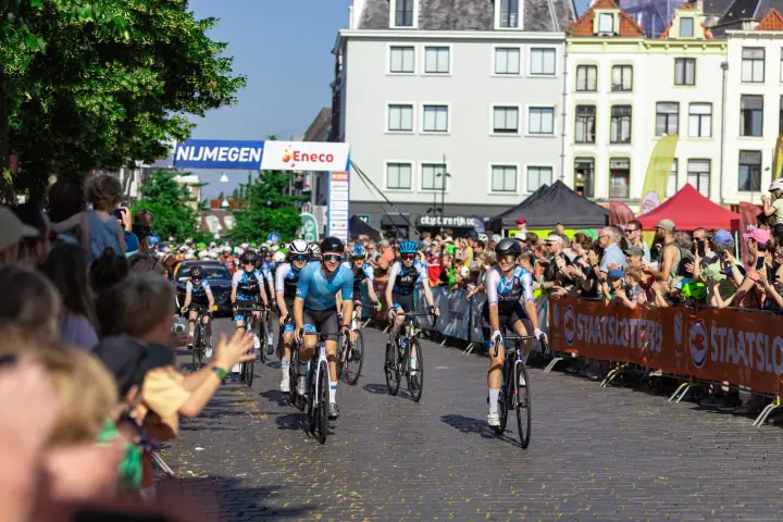 Groep renners rijdt over de kasseien onder de startboog van Nijmegen weg, langs volle dranghekken