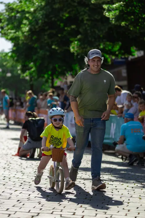 Kind met een mandje aan de loopfiets, met een lachende begeleider die naast hem meeloopt