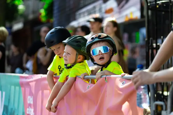 Drie kinderen met fietshelm hangen over het dranghek om de kinderrace te volgen