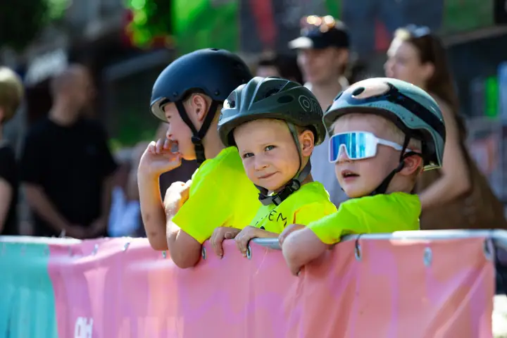 Jongen met groene helm kijkt lachend in de camera, naast twee andere kinderen aan het hek