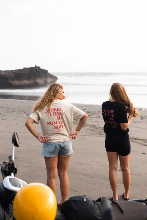 Twee vrouwen met de handen in de zij kijken uit over het strand, een gele helm en scooter vooraan.