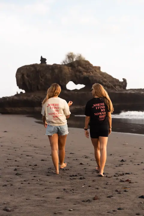 Twee vrouwen in het beige en zwarte Nomadwise shirt lopen over het strand naar een rotsboog.