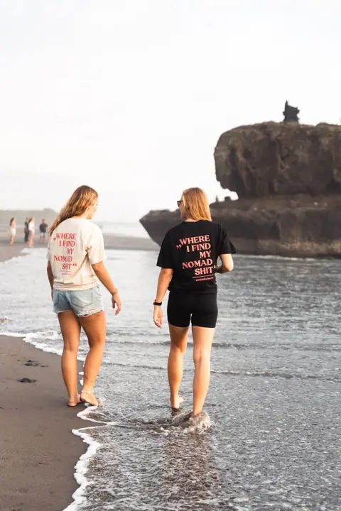 Blootsvoets door de branding, twee vrouwen in Nomadwise shirts met een rotsformatie ernaast.