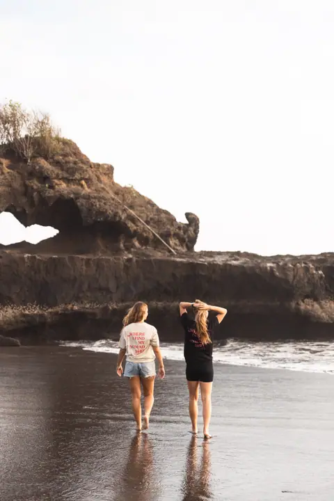 Op het natte, donkere zand lopen twee vrouwen weg van de camera, richting een rots met een gat erin.