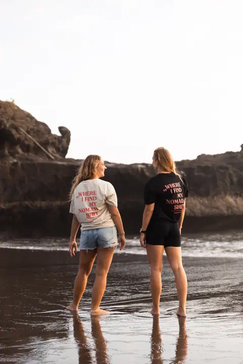 Twee vrouwen staan op blote voeten op het spiegelende zand en kijken elkaar aan, rotsen erachter.