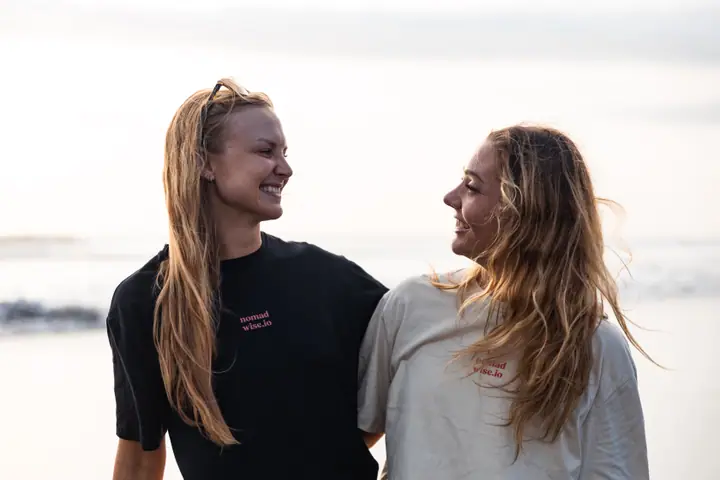 Twee vrouwen lachen elkaar toe aan zee, op hun borst het kleine nomadwise.io logo.
