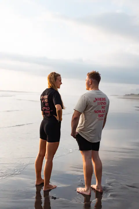 Een man en een vrouw staan blootsvoets op het natte zand en kijken elkaar aan, beide rugprints zichtbaar.