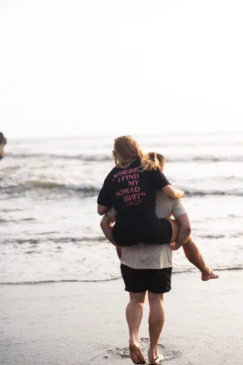 Van achteren gezien loopt een man met een vrouw op zijn rug de golven in.