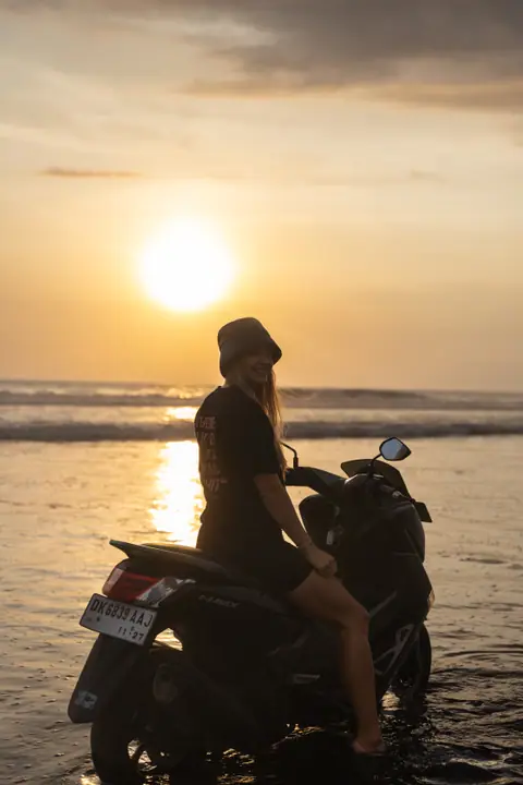Vrouw op een scooter in het ondiepe water kijkt lachend om, met de laagstaande zon achter haar.