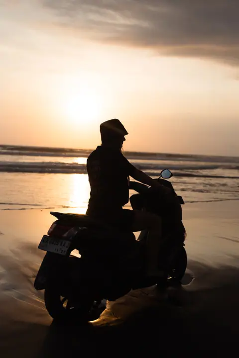 Silhouet van iemand met een hoedje op een scooter, tegen de ondergaande zon boven zee.