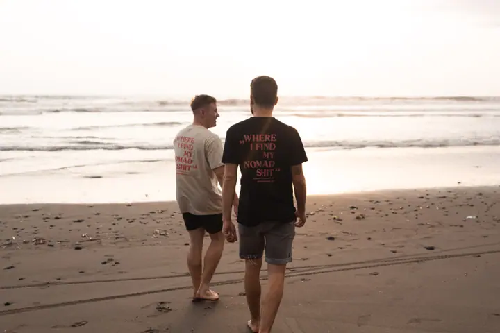 Twee mannen in een zwart en een beige shirt lopen samen over het strand richting zee.