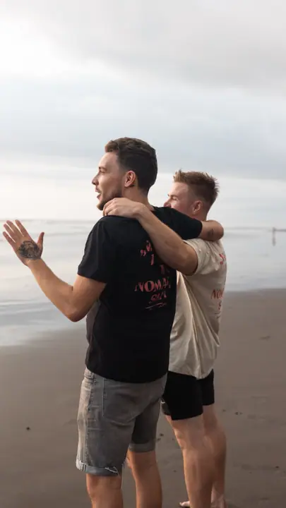 Arm in arm op het strand, een van de mannen gebaart lachend naar zee.