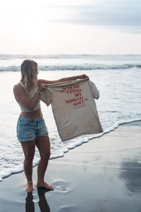Een vrouw houdt in de branding het beige Nomadwise shirt omhoog om de rugprint te laten zien.