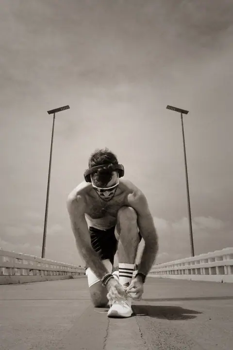 Lukas van Run Freak strikt zijn veters op een lege brug, met koptelefoon en sportbril