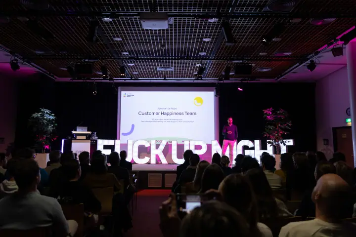 Overzicht van de volle zaal met een spreker op het podium tussen het scherm en de verlichte F*ckup Night letters