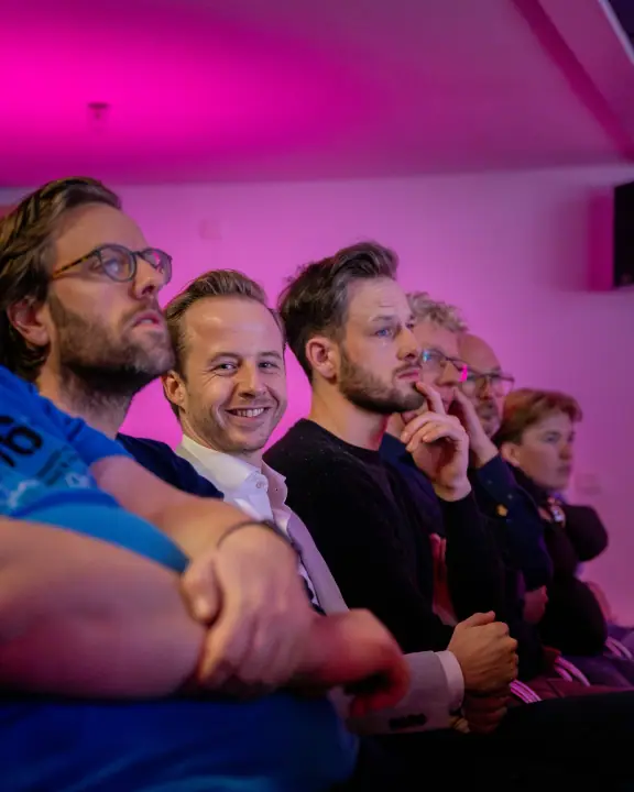 Rij bezoekers in roze licht, een man in wit overhemd lacht recht in de camera