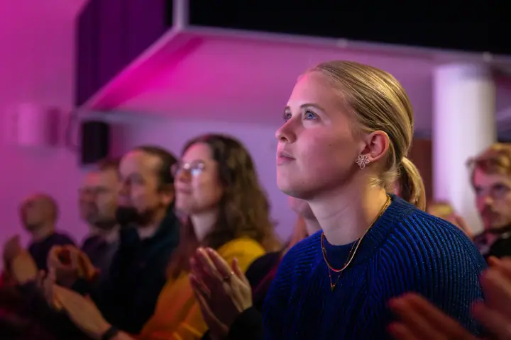 Jonge vrouw in blauwe trui applaudisseert en kijkt omhoog naar het podium