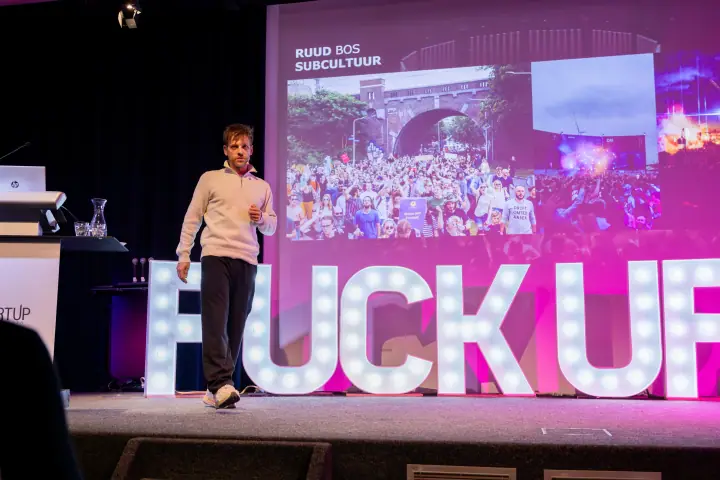 Spreker wandelt in sneakers over het podium, vlak voor de witte lichtletters van de avond