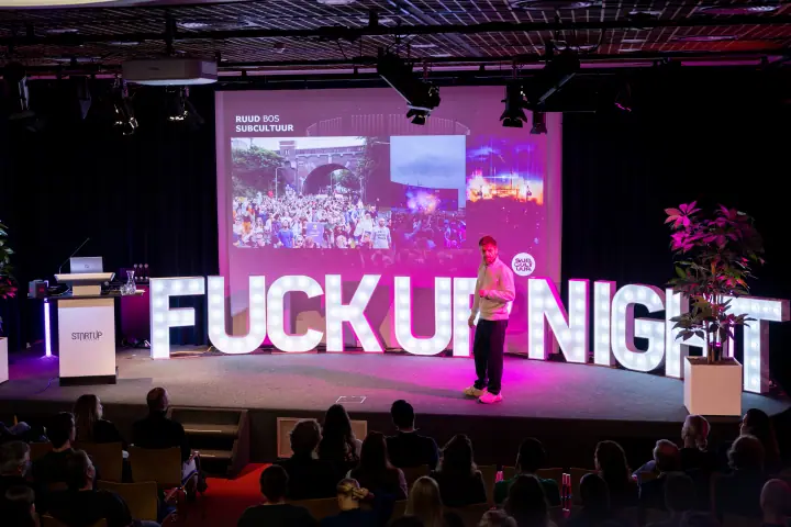 Podium van de Startup F*ckup Night met spreker, planten, katheder en lichtletters, gezien vanuit de zaal
