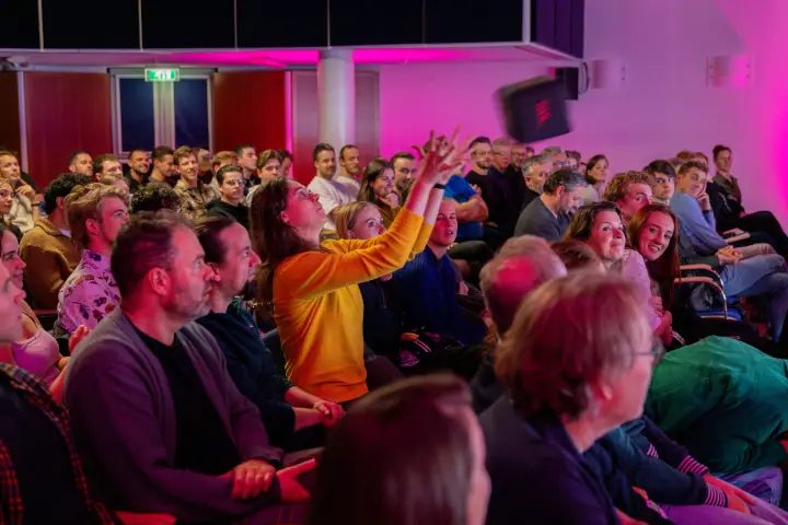 Vrouw in oranje trui steekt haar armen in de lucht in een lachende, volle zaal