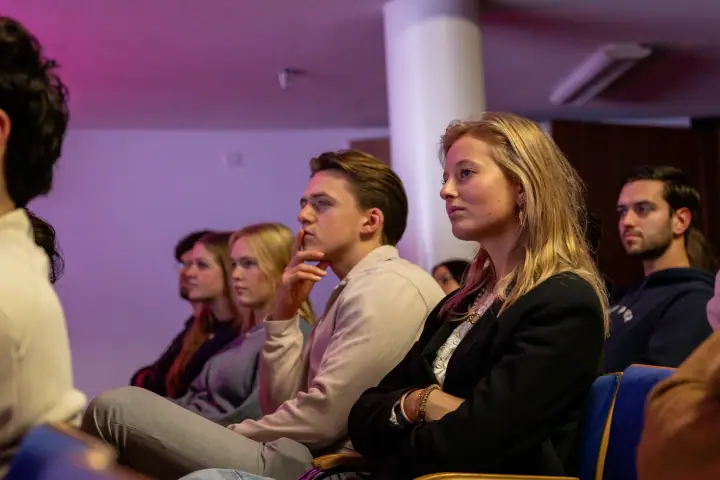 Jonge vrouw in zwart jasje en een jonge man met zijn hand aan zijn kin luisteren naar de spreker