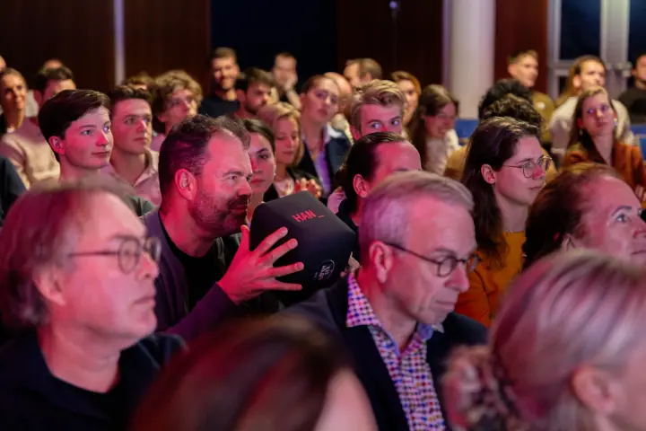 Bebaarde man in het publiek spreekt in de zachte gooimicrofoon, omringd door luisterende bezoekers