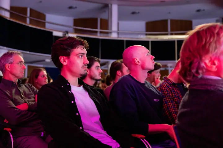Bezoekers in donkere kleding volgen de talk met serieuze blikken, verlicht door het podiumlicht