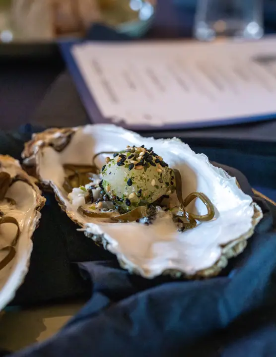 Bolletje met sesamzaad op zeewier in een oesterschelp, met de menukaart onscherp erachter