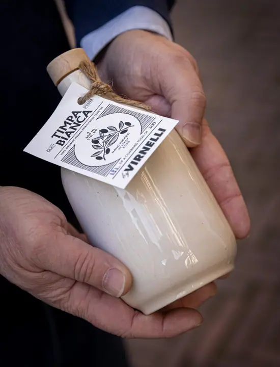 Crèmekleurige stenen fles Timpa Bianca met touwtje en label, rustend in twee handen