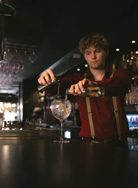 Bartender met krullen en bretels schenkt uit een jigger en een flesje tonic in een ballonglas