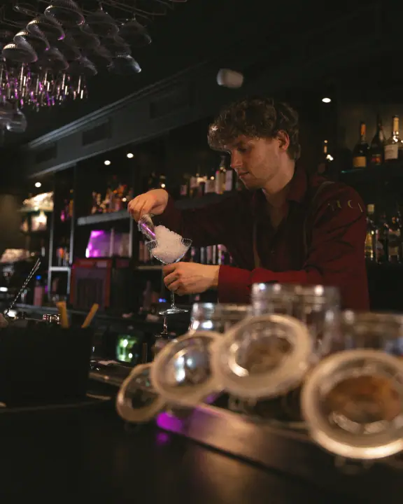 Bartender vult een glas met crushed ijs, vooraan op de bar een rij weckpotten