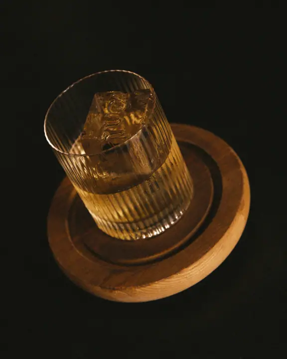 Geribbeld tumblerglas met een helder ijsblok met ingestempeld logo op een houten onderzetter