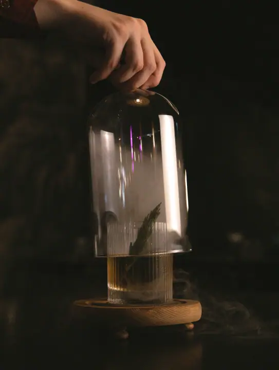 Een hand tilt de glazen stolp op, waardoor een gerookte cocktail tevoorschijn komt