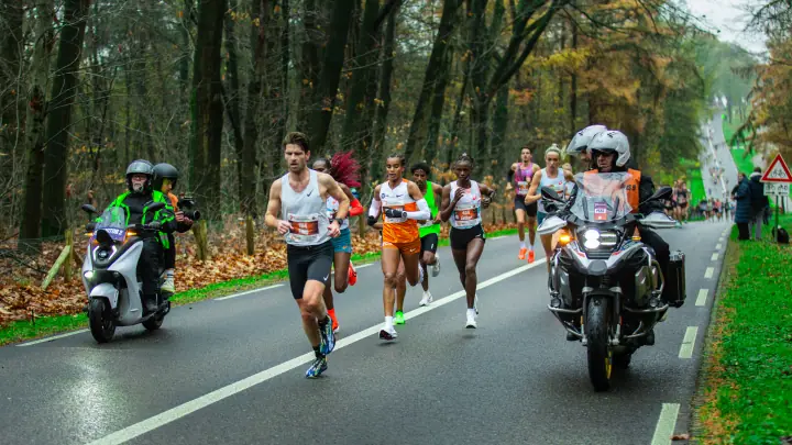 Kopgroep van de Zevenheuvelenloop 2025 rent door het herfstbos, geflankeerd door twee motoren